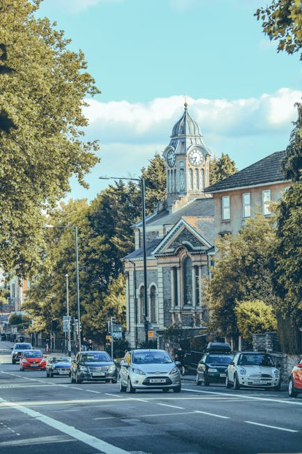 A row of Victorian-style terraced houses in Dulwich with ornate ironwork balconies, large bay windows, and decorative brickwork, set against a bright blue sky with some scattered clouds. The houses ar