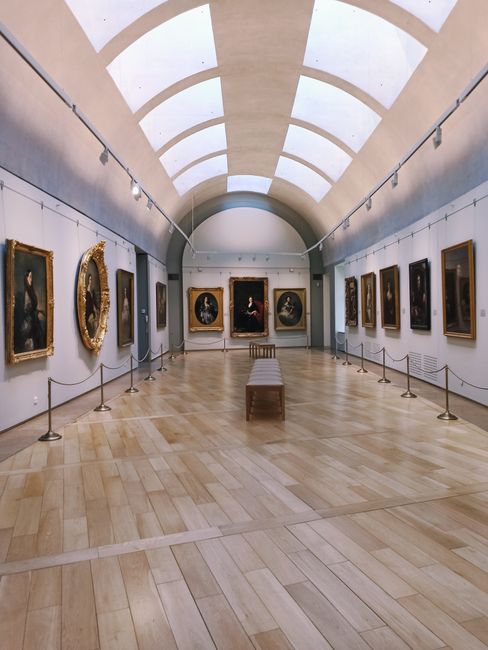 An interior view of Dulwich Picture Gallery with a curved, white, vaulted ceiling featuring large skylights that allow natural light to illuminate the space. The gallery walls are lined with framed cl
