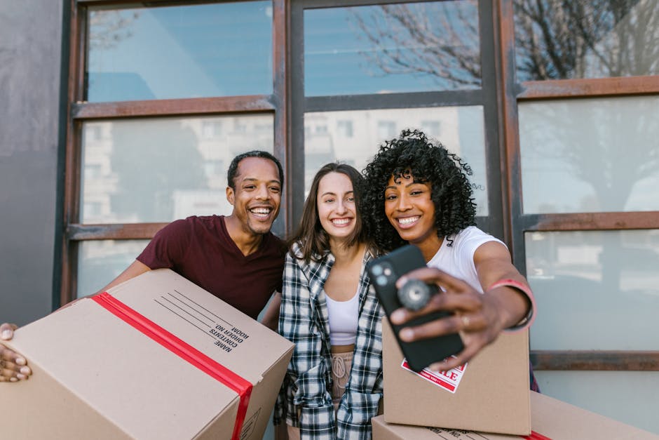 Three smiling individuals standing outside a building with large windows, holding cardboard boxes sealed with red tape, during a home relocation process. The person on the right is taking a selfie with a smartphone, capturing the group and the boxes. The boxes are made of cardboard, with one displaying a printed label. They are dressed casually, and the environment suggests a bright day with natural light. This scene illustrates the packing and moving preparations typical of house removals services, with visible handling of boxes and coordination for transportation purposes, supported by Man with Van Dulwich in their professional removals service for West Dulwich station relocations, DULWICH.