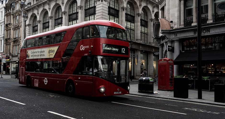 A red double-decker London bus marked with route number 12 and destination 'Dulwich Library' is parked on a busy street in West Dulwich, London, with a shopfront and a traditional red telephone booth visible on the sidewalk. The scene includes black pavement with white markings, metal trash bins, and a lamppost, all set against a backdrop of historic multi-story buildings with large windows and ornate architecture. The bus's interior windows and exterior signage are visible. This image depicts urban transportation as part of a home relocation or moving service context, with potential use for illustrating furniture transport, packing, and moving logistics in the Dulwich area. Man with Van Dulwich, specializing in removals, might utilize such imagery to represent their local moving and house removal services.