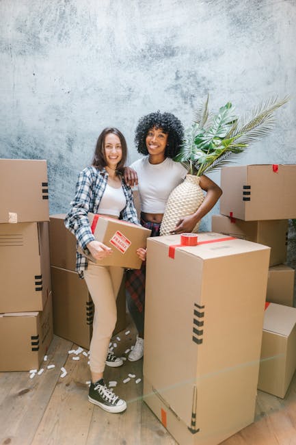 Three smiling individuals standing outside a building with large windows, holding cardboard boxes sealed with red tape, during a home relocation process. The person on the right is taking a selfie with a smartphone, capturing the group and the boxes. The boxes are made of cardboard, with one displaying a printed label. They are dressed casually, and the environment suggests a bright day with natural light. This scene illustrates the packing and moving preparations typical of house removals services, with visible handling of boxes and coordination for transportation purposes, supported by Man with Van Dulwich in their professional removals service for West Dulwich station relocations, DULWICH.